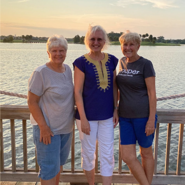 Three women standing on a pier in front of a lake, smiling.